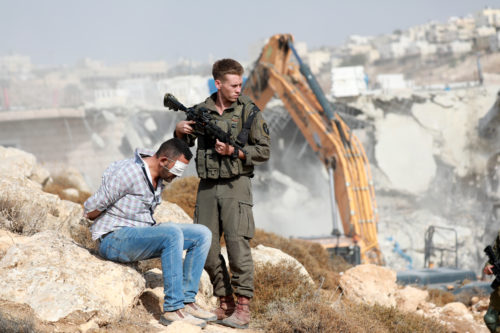 Israelilaissotilas seisoo palestiinalaisen miehen vieressä. Mies istuu silmät sidottuina.