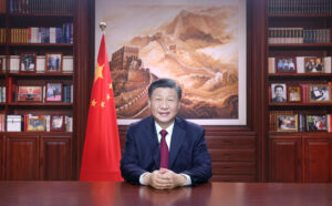 Xi Jinping istuu pöydän ääressä takanaan kirjahylly, jossa on kuvia ja kirjoja sekä Kiinan lippu ja maalaus.