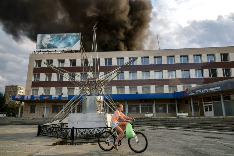 Tyttö pyöräilemässä ohjusten osia valmistavan tehtaan edessä. Tehtaan katolta nousee savua.